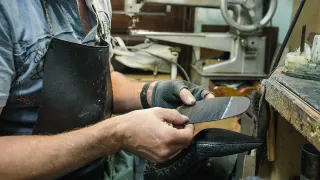 Manos de un zapatero con mandil de cuero ajustando una suela contra un zapato negro, con una máquina de coser de poste al fondo del taller