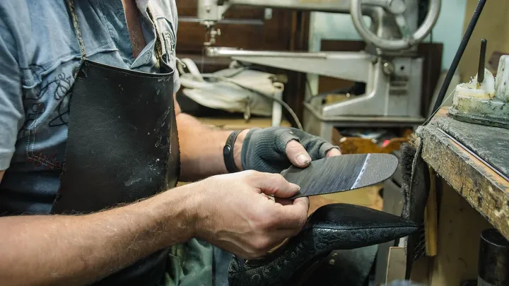 Manos de un zapatero con mandil de cuero ajustando una suela contra un zapato negro, con una máquina de coser de poste al fondo del taller