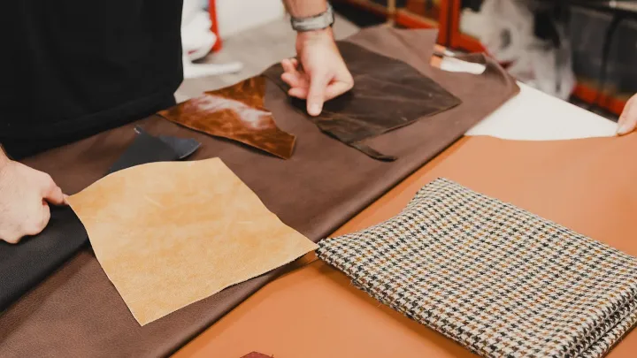 Manos sobre una mesa de taller con recortes de cuero de calidades distintas: uno claro con flor visible, uno engrasado brillante, uno oscuro envejecido y uno negro, junto a una tela de pata de gallo