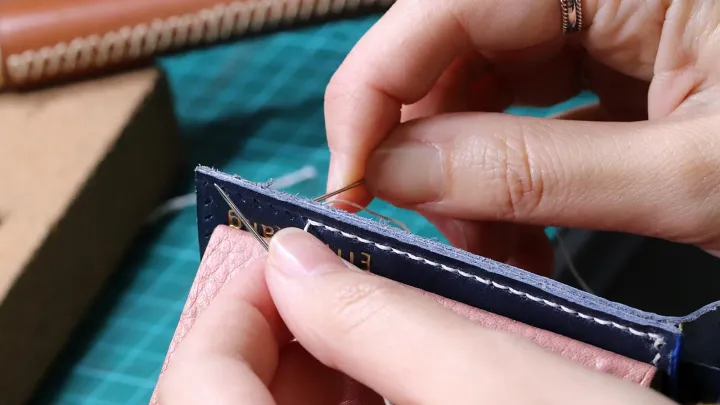 Manos cosiendo cuero a mano con aguja e hilo encerado blanco, con la costura recta a medio hacer en el borde de una pieza azul