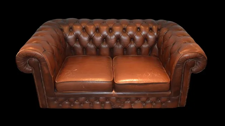 Sofá chesterfield de dos plazas con tapizado de cuero color coñac, capitoné con botones, tachuelas en los brazos y la pátina del uso en los cojines
