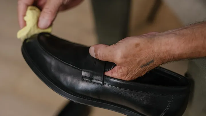 Manos limpiando con un paño suave amarillo un mocasín de cuero liso negro, el paso básico para limpiar cuero con acabado