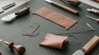 Mesa de marroquinería con dos piezas de billetera en cuero café, una a medio coser con hilo encerado, sobre una plancha de corte rodeada de cuchillas y una lezna