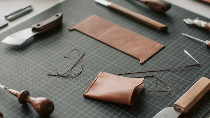 Mesa de marroquinería con dos piezas de billetera en cuero café, una a medio coser con hilo encerado, sobre una plancha de corte rodeada de cuchillas y una lezna