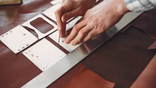 Manos marcando con un punzón el contorno de moldes de cartulina cuadriculada para billetera, uno con la ventana del tarjetero, sobre cuero café junto a una regla metálica