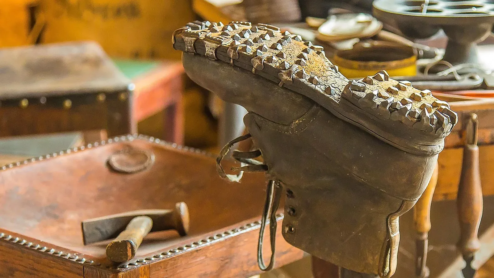 Bota de cuero gastada con suela claveteada, montada boca arriba sobre una horma de hierro, con un martillo de zapatero sobre la mesa del taller
