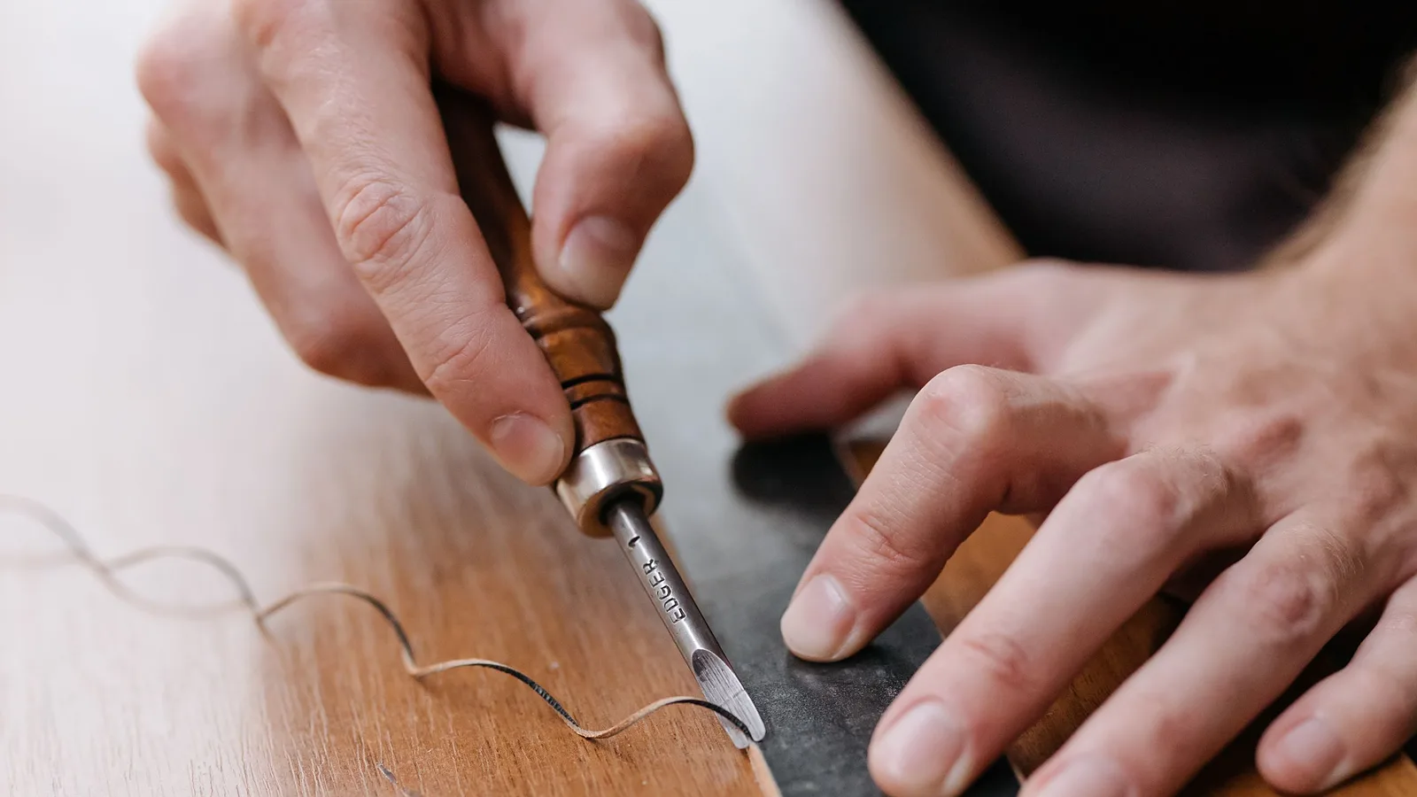 Manos de artesano canteando el borde de una tira de cuero con una herramienta de mango de madera sobre la mesa del taller