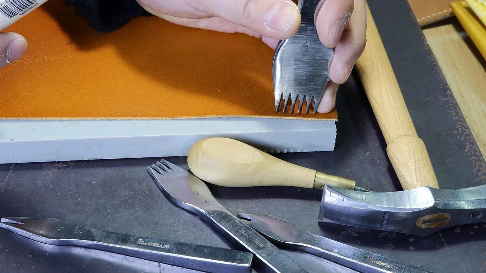 Manos marcando puntadas con una pinza de coser sobre una pieza de cuero color naranja en un banco de trabajo, con mazo y otras pinzas alrededor
