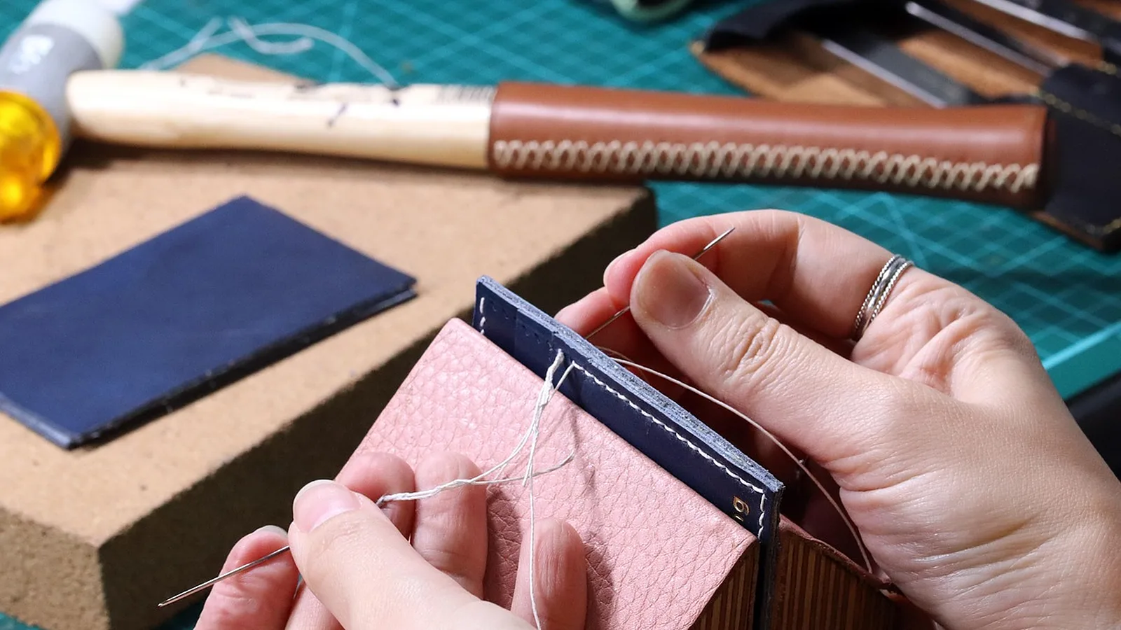 Manos cosiendo a mano con punto silla, una aguja en cada extremo del hilo, una billetera pequeña de cuero rosa y azul, con un mazo y la plancha de corte al fondo