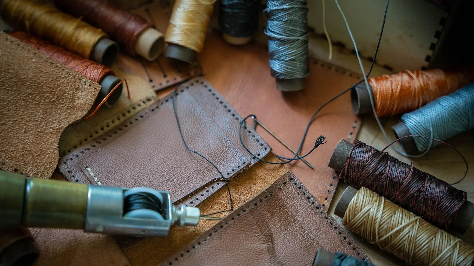 Carretes de hilo encerado de varios colores sobre piezas de cuero café ya perforadas, con una aguja enhebrada y una lezna de coser